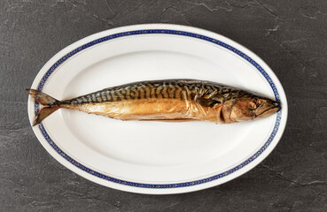 Whole smoked mackerel fish on white oval plate, gray black stone like table under. View from above