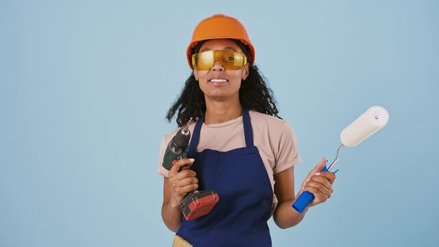 Dark-skinned lady in hard hat and protective goggles. Holding paint roller, smiling, acting like shooting you with drill. Posing on blue background
