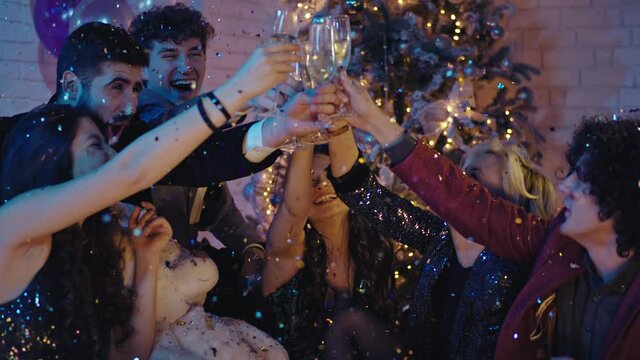 Beside the Christmas tree group of friends celebrating together louts of confetti all over them they cheers with glasses of champagne