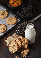 Homemade organic oatmeal cookies with raisins and apricots with baking tray and bottle of milk on grey wooden background. Black bowl of raisins and dried apricots.Top view