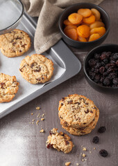 Homemade organic oatmeal cookies with raisins and apricots with baking tray on grey wooden background. Black bowl of raisins and dried apricots.Top view