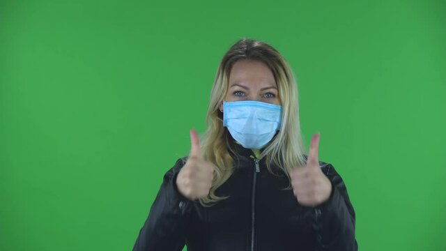 Portrait of beautiful young woman in medical protective face mask looking at camera and showing thumbs up gesture with both hands. Blonde with loose hair in a black jacket and jeans on a green screen