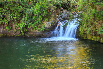 Raparapahoe Falls in the Bay of Plenty, New Zealand, cascading into a pool surrounded by native forest. Two men are climbing up the waterfall with the help of a rope