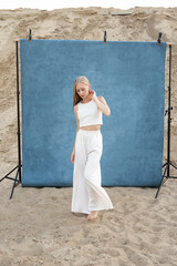 Obraz premium beauty portrait outdoors on sand in front of blue studio background, pretty female posing. attractive caucasian teen girl with long hair. beautiful young woman in white clothes, elegant fashion style