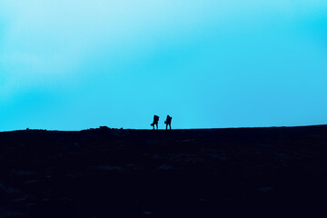 Fototapeta premium Travelers walk along a trail in the mountains. Silhouettes of two adventure hikers with backpacks walk in the mountains at dusk.