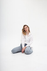 portrait of young pensive caucasian woman posing in shirt and blue jeans, sitting on white studio floor on her knees. model tests of pretty girl in basic clothes on cyclorama. attractive female poses