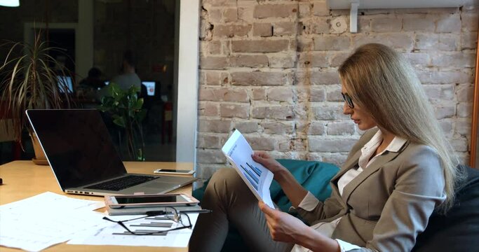 Close-up portrait of the beautiful blonde businesswoman wearing glasses is working with the documents while sitting at the laptop in the office.