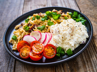 Delicious breakfast - scrambled eggs with fried sausages, cottage cheese and vegetables served on black plate on wooden table
