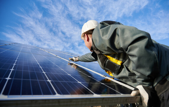 Side view worker wearing uniform, working gloves and helmet under blue sky on background. Technician setting a shiny new solar battery with help of hex key. Concept of alternative green energy