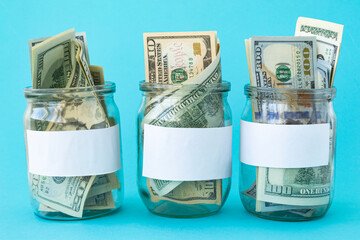 Three Piggy Bank from a Glass Jar for Life. New dollars sticking out of piggy banks on a blue background. The banks have white sheets for inscriptions. Mockup. Copy space. Economy concept