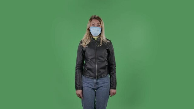 Portrait of beautiful young woman in medical protective face mask looking at camera worries and scared. Blonde with loose hair in a black jacket and jeans on a green screen in the studio. Health