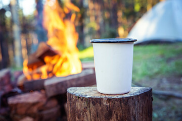Close up of white camping cup, on top of wood stump, in front of campfire and sleeping tent.