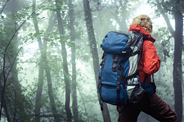 Mountain hiking woman, wearing backpack and outdoor clothes, walking through deep, misty forest, with available copy space next to her.