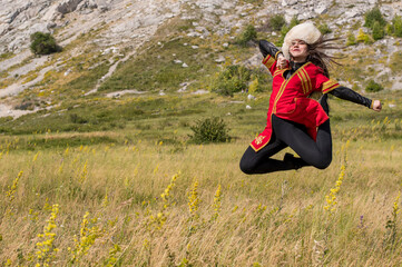 A cheerful young girl in a red suit soars over a green field, dancing a national dance on a mountain background
