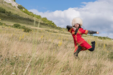 Naklejka na ściany i meble A young cheerful girl in a national costume is dancing a fiery dance on a green field at the foot of the mountain
