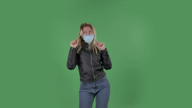 Portrait of beautiful young woman in medical mask is looking straight and is dancing funny. Blonde with loose hair in a black jacket and jeans on a green screen in the studio. Health Protection Corona