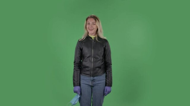 Portrait of beautiful young woman which is removes medical protective mask breathes air and smiling. Blonde with loose hair in a black jacket and jeans on a green screen in the studio. Health