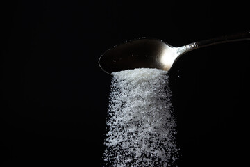 Naklejka na ściany i meble sugar is poured from a spoon on a black background. Excessive sugar intake. Sweet crystals