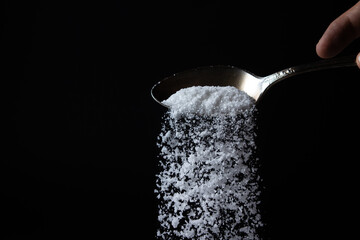 Naklejka na ściany i meble Salt is poured from a spoon on a black background. Excessive salt intake. Coarse salt