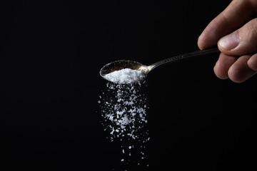 Naklejka na ściany i meble Salt is poured from a spoon on a black background. Excessive salt intake. Coarse salt
