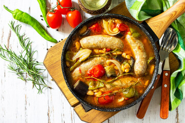 Stew of beans, vegetables and smoked sausages with tomato sauce in the pan on wooden table. Top view flat lay background.