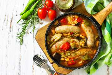 Stew of beans, vegetables and smoked sausages with tomato sauce in the pan on wooden table. Top view flat lay background. Copy space.