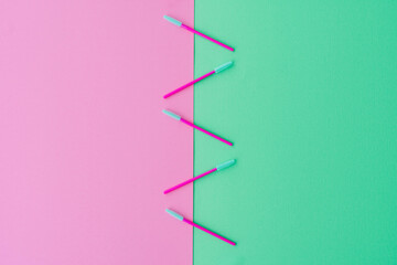 Beauty mascara wands in pink and teal green arranged in a line through the centre of the frame.  Pink and green background with copy space.  Flat lay style beauty business background.