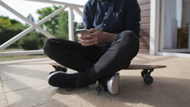 Young man using a smartphone while sitting relaxed at longboard on terrace of house, close up