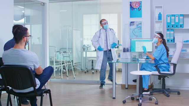 Nurse with visor against coronavirus in hospital waiting area scanning senior man forehead for temperature using infrared thermometer. Doctor with patient in cabinet. Invalid woman in wheelchair.