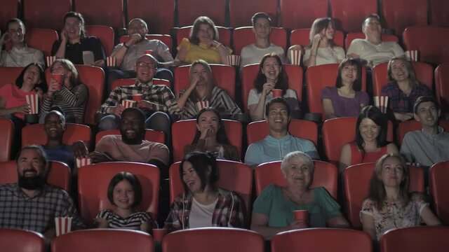 Joyful multi-ethnic spectators laughing while watching comedy movie in cinema. Diverse people having fun, eating popcorn, drinking soda in movie theater, film industry and entertainment concept