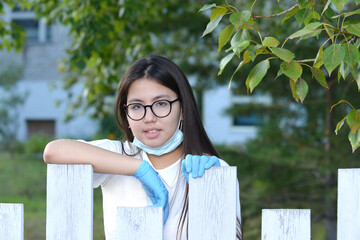 A schoolgirl wearing medical gloves and a mask stands behind a fence in the background of a school. A girl in protective equipment against the background of green trees.