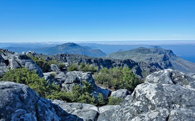 Fototapeta na wymiar Alpine landscape. Ancient gray boulders lie on the flat summit of Table Mountain. Low shrubs, fynbos grow between the stones. In the distance, mountain peaks hide in the clouds. Cape Town.South Africa
