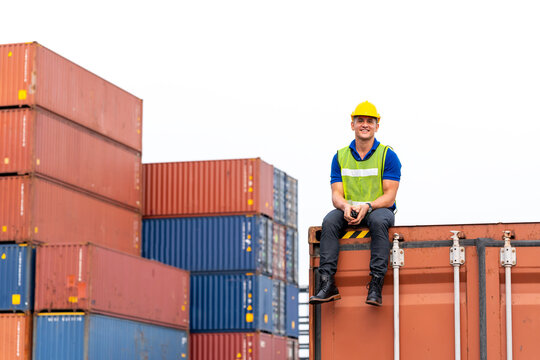 Portrait of confident inspector sitting on cargo container in shipping yard, Smiley engineer or happy dock worker wears hardhat and safety vest in port. Concept of transportation, logistics business.
