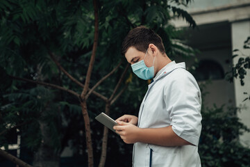 Doctor standing outside and holding tablet pc. Medical staff with surgical face mask against corona virus. Male nurse wearing white gown and stethoscope to work with patients. Copy space.