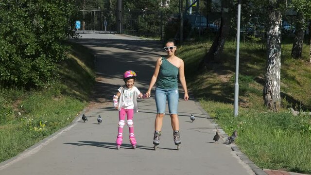 Happy Young Mother and her Cute Little Daughter Inline Skating Together in a City Park. Slow Motion. Childhood, Summer Activities and Mothers Day Concept
