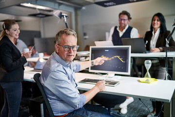 Portrait of confident mature businessman pointing at graph on computer screen while working with colleagues in creative workplace