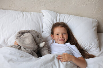 Cute child little girl wake up and lies in the bed with a toy elephant.