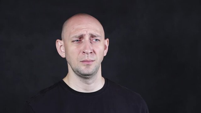 A bald caucasian man in a black t-shirt on a black background depicts a grimace of discontent close-up. High quality FullHD footage