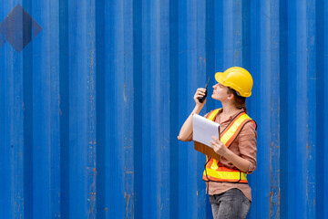 Confident inspector standing in front of cargo container in shipping yard. Smart female engineer, dock worker wear hard hat using clipboard and radio to command transportation. Concept of logistics