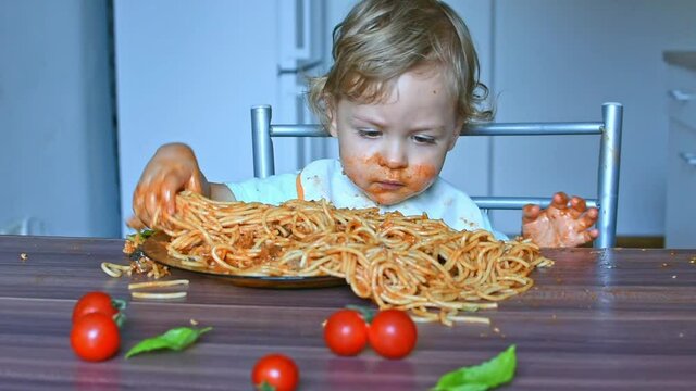 Happy sweet baby boy making a mess in the kitchen while eating spaghetti. Funny toddler child with home made pasta and tomato sauce
