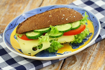 Whole grain roll with Swiss cheese, lettuce, tomato and cucumber on colorful vintage plate on wooden surface, closeup
