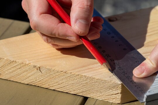 A closeup portrait of a man using a pencil and a stainless steel L-square or snag to draw a perpendicular line across a wooden plank.