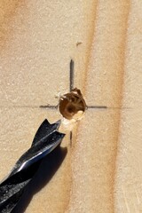 A closeup portrait of a wooden plank with a hole drilled into it on the intersection of the lines of a pencil marking with the wood drilling bit lying next to it.