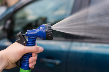 A woman's hand holds a hose for washing the car. Water, spray, jet. The idea is to wash your car in front of your house, saving after a pandemic. Photo close-up, horizontal.