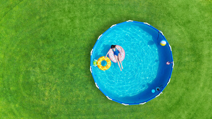 Aerial. Girl swims in a metal frame pool with inflatable toys. Frame pool stand on a green grass lawn. Top view. Copy space. - obrazy, fototapety, plakaty