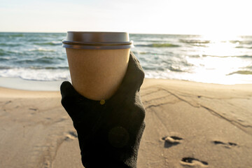 Coffee at the sea. Young woman with gloves holds a disposable cup of coffee on on a sunny morning at the beach. 