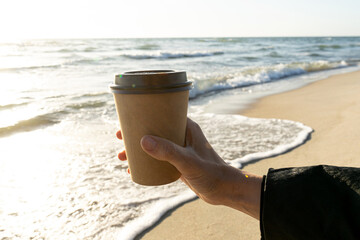 Coffee at the sea. Young woman holds a disposable cup of coffee on on a sunny morning at the beach. 