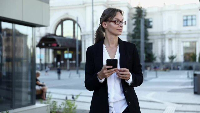 Beautiful woman in formal clothing checking arrival time of public transport on smartphone. Business lady with brown hair walking on bus stop and using modern gadget.