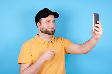 Brunette man in black hat using mobile phone, makes selfie. Isolated on blue background.