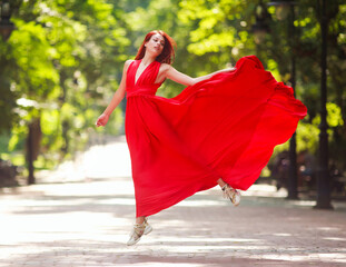 Young woman in luxury long red dress dancing, jumping in city park